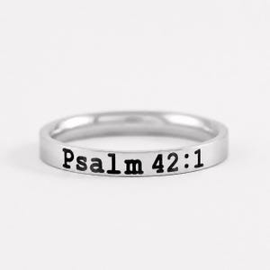 May include: A silver-toned metal band ring featuring a smooth, polished finish. The exterior surface of the ring is engraved with the black text Psalm 42:1, which is clearly visible and centered along the front portion of the circular band.