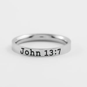 May include: A silver-colored ring with the text "John 13:7" in black lettering. The ring is circular and appears to be made of metal. The background is a solid, light color.