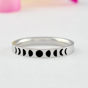 May include: A silver-toned metal ring featuring a series of black moon phase icons engraved along the outer band. The design displays the lunar cycle from a thin crescent to a full moon and back to a crescent, set against a polished metallic surface.