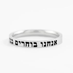 May include: A polished silver-toned band ring featuring a flat outer surface engraved with black Hebrew text that reads "אנחנו בוחרים בנו". The ring has a smooth, metallic finish and a consistent, slim width throughout its circular design.