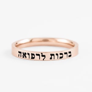 May include: A rose gold-toned band ring featuring a flat outer surface engraved with black Hebrew text that reads "ברכות לרפואה". The ring has a smooth, polished finish and a consistent width throughout its circular design, showcasing a simple metal construction.