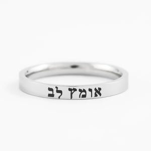 May include: A silver ring with the Hebrew text "אומץ לב" engraved in black. The ring has a smooth, polished surface and a simple, elegant design. The ring is likely made of stainless steel or a similar metal.