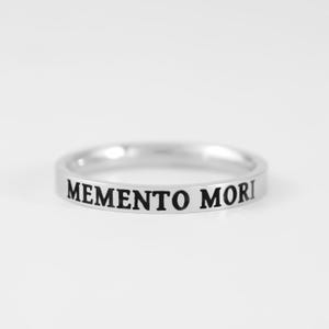 May include: A silver-toned metal ring featuring a smooth, polished finish. The outer surface of the band is engraved with the black-filled text MEMENTO MORI in a serif font, encircling the circumference of the ring in a simple, clean design.