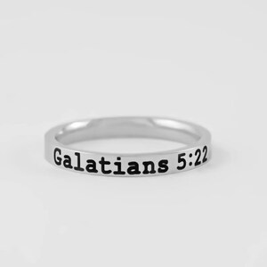 May include: A silver-toned metal band ring featuring the black engraved text Galatians 5:22 along the outer surface. The ring has a smooth, polished finish and a slim, uniform width, presenting a clean and simple design for everyday wear.