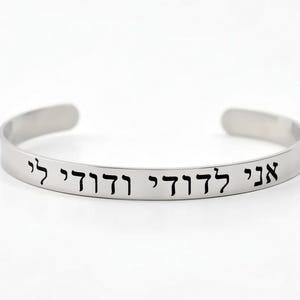 May include: A silver-colored cuff bracelet with Hebrew text engraved in black. The bracelet has a simple, open design and is displayed against a white background. The text is clearly visible.