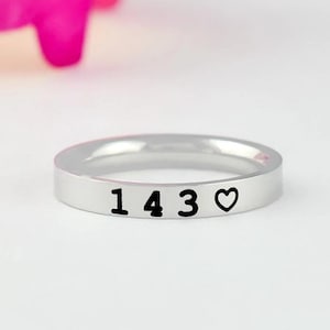 May include: A silver-toned metal ring with a smooth, polished finish. The outer surface of the band features the black engraved numbers 1 4 3 followed by a small black heart symbol. The ring has a simple, flat, and uniform band design.