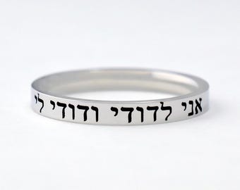 Könnte beinhalten: Ein polierter silberfarbener Bandring mit einer glatten, flachen Oberfläche. Die Außenseite des Rings ist mit dem schwarzen hebräischen Text "אני לדודי ודודי לי" graviert. Der Ring hat eine schmale, gleichmäßige Breite und eine reflektierende Oberfläche.