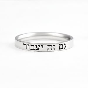 May include: A polished silver-tone band ring featuring the Hebrew inscription "גם זה יעבור" engraved in black lettering along the outer surface. The ring has a smooth, flat profile and a consistent width, showcasing a clean and simple design.