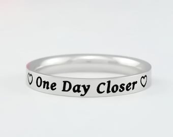 May include: A silver-toned metal ring featuring the engraved black text "One Day Closer" centered on the outer band, flanked by a small heart symbol on each side of the phrase. The ring has a smooth, polished finish and a simple band design.
