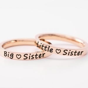 May include: Two rose gold rings, one reading "Big Sister" with a heart symbol, and the other reading "Little Sister" with a heart symbol. The rings are set against a white background.