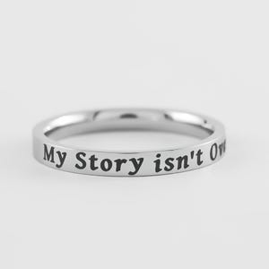 May include: A silver ring with the engraved text "My Story isn't Over". The ring is circular and has a polished, reflective surface. The text is in a bold, sans-serif font.