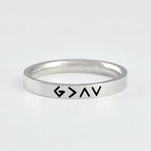 May include: A polished silver-toned metal band ring featuring a flat outer surface. The ring is engraved with the black text G > ^ V, which represents the phrase God is greater than the highs and lows. The band has a smooth, reflective finish.