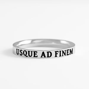 May include: A polished silver-toned band ring featuring the engraved black text USQUE AD FINEM centered on the outer surface. The ring has a smooth, rounded profile and a reflective metallic finish, showcasing clear, bold lettering in a serif font.