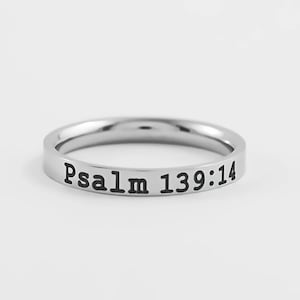 May include: A silver-colored ring with the text "Psalm 139:14" engraved in black. The ring is simple and has a smooth, polished finish. The ring is a piece of jewelry.
