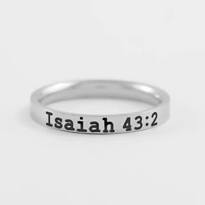 May include: A silver-toned metal band ring featuring the engraved black text Isaiah 43:2 on the outer surface. The ring has a smooth, polished finish and a simple, uniform width throughout the entire circumference of the circular band.