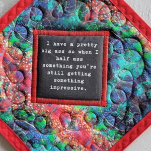 Puede incluir: Manopla acolchada en forma de diamante con un patrón vibrante en tonos azules, verdes y rojos. Tiene un borde rojo y un lazo para colgar. Un cuadrado negro en el centro presenta el texto: "I have a pretty big ass so when I half ass something you're still getting something impressive."