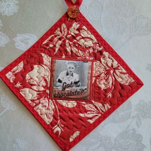 Puede incluir: Manopla de cocina roja acolchada con un patrón floral blanco. Una foto cuadrada de una persona con un gorro de cocinero está unida a la manopla de cocina. La foto tiene el texto "Got chocolate?" impreso en ella.
