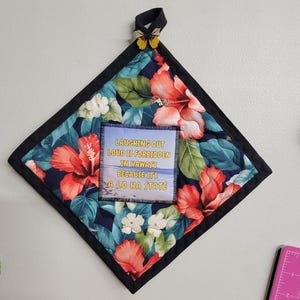 Puede incluir: Un salvamanteles cuadrado acolchado con un borde negro, con un estampado floral tropical con flores de hibisco rojas y hojas verdes. El centro tiene una imagen impresa con el texto "LAUGHING OUT LOUD IS FORBIDDEN IN HAWAII BECAUSE ITS A LO HA STATE". Un lazo negro y un adorno de mariposa en la parte superior.