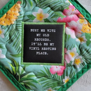 May include: A green and white tropical print pot holder with a black square patch that reads "BURY ME WITH MY OLD RECORDS. IT'LL BE MY VINYL RESTING PLACE."