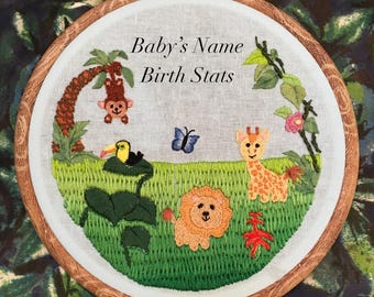 May include: Embroidered hoop art with a jungle theme. It features a toucan, a monkey, a giraffe, a lion, and a butterfly. The hoop has a white background with the text "Baby's Name Birth Stats" in black.