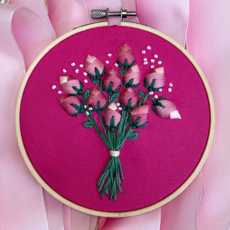 May include: Embroidered hoop art featuring a bouquet of pink roses with green stems and leaves on a bright pink background.