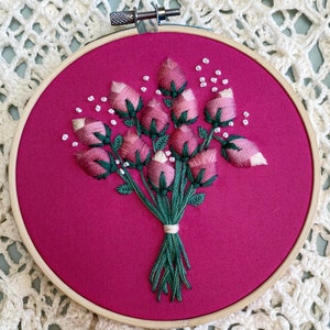 May include: A pink and green embroidered bouquet of flowers on a pink fabric background. The flowers are arranged in a circle and are surrounded by white beads.