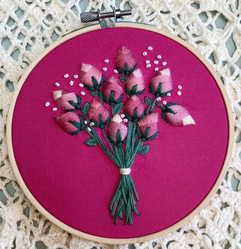 May include: A pink and green embroidered bouquet of flowers on a pink fabric background. The flowers are arranged in a circle and are surrounded by white beads.