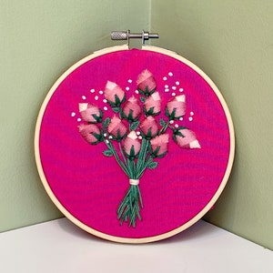 May include: A pink and green embroidered bouquet of roses in a wooden embroidery hoop.