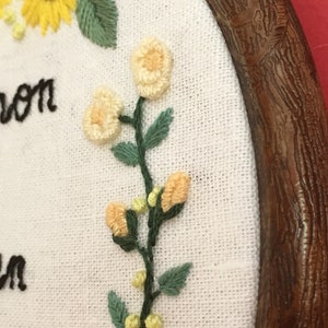May include: Close-up of a white fabric with embroidered yellow sunflowers, white flowers, and green leaves. The fabric is framed in a brown wooden hoop. The text "Alison" is embroidered on the fabric.