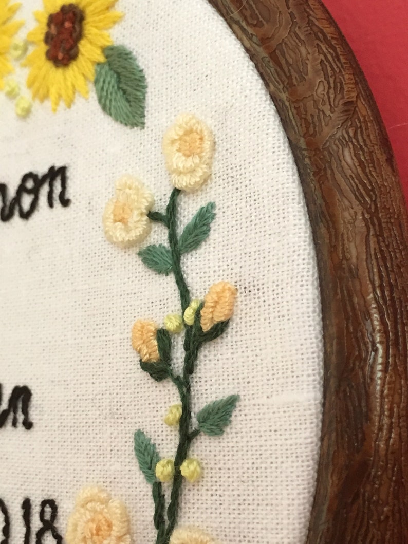 May include: Close-up of a white fabric with embroidered yellow sunflowers, white flowers, and green leaves. The fabric is framed in a brown wooden hoop. The text "Alison" is embroidered on the fabric.