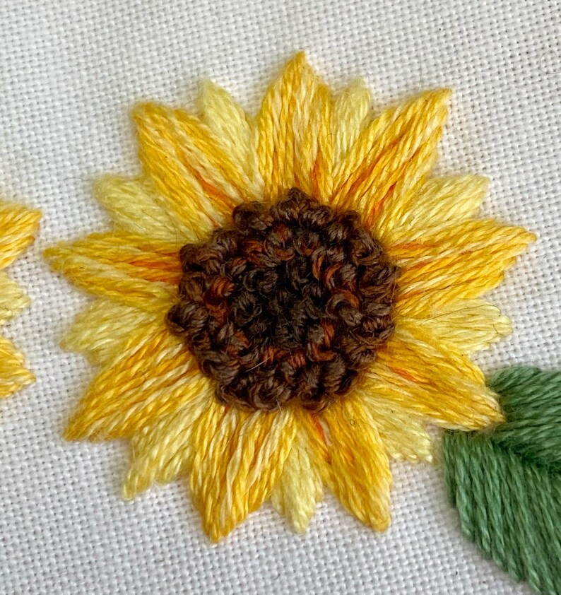 May include: Close-up of an embroidered sunflower with a brown center and yellow petals. The sunflower is stitched on a white fabric background.
