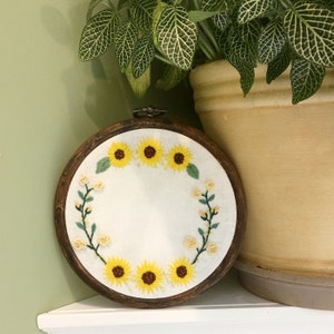 May include: A wooden embroidery hoop with a white fabric background featuring a wreath of embroidered sunflowers and small white flowers.