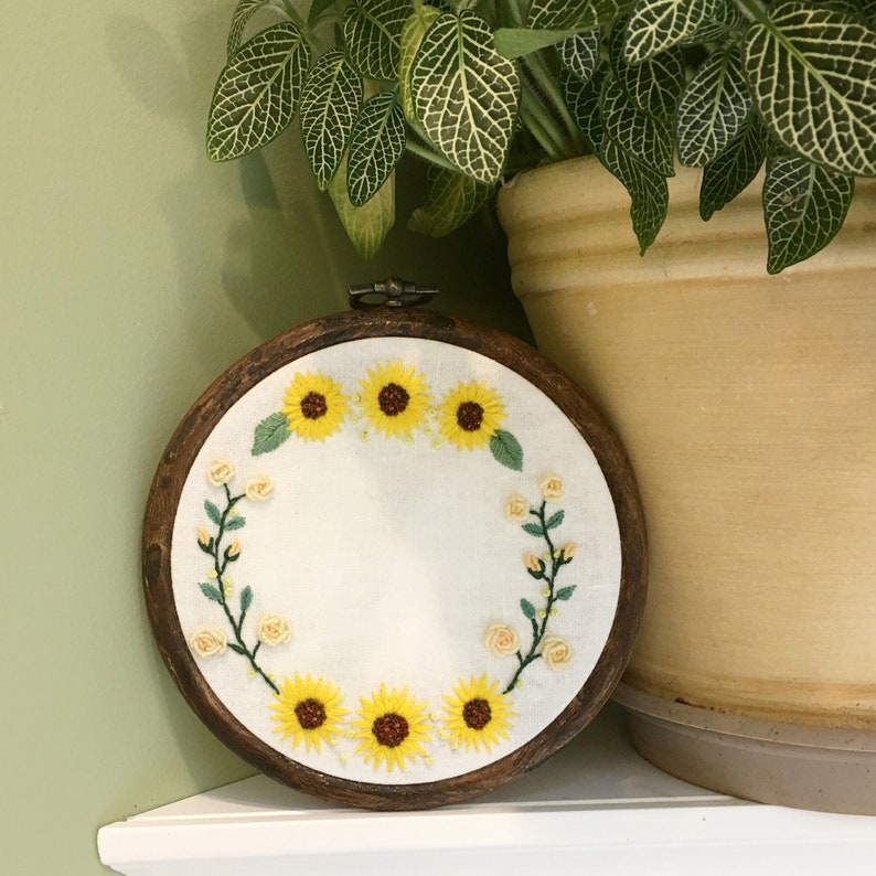May include: A wooden embroidery hoop with a white fabric background featuring a wreath of embroidered sunflowers and small white flowers.