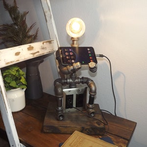 May include: A steampunk-style lamp made from pipes and fittings, with a light bulb and a phone holder. The lamp is sitting on a wooden table with a couple of books.