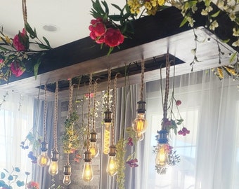 May include: A rustic wooden chandelier with multiple light bulbs hanging from rope cords. The chandelier is decorated with artificial flowers in shades of pink, white, and yellow.