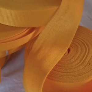 May include: Multiple rolls of bright yellow webbing tape featuring a dense, woven texture. The material is wound into tight, circular coils, with one end of the tape extending outward to display the flat, durable surface of the fabric ribbon.
