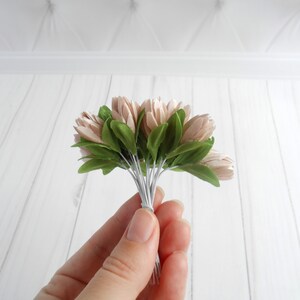 May include: A small bouquet of artificial flowers held in a hand. The flowers are a soft pink color with green leaves. The stems are wrapped together with white wire. The background is a white wooden surface.