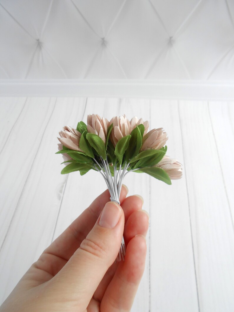 May include: A small bouquet of artificial flowers held in a hand. The flowers are a soft pink color with green leaves. The stems are wrapped together with white wire. The background is a white wooden surface.