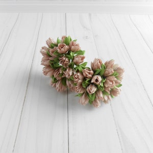 May include: Two clusters of artificial flowers with light pink petals and green leaves. The flowers are arranged in a round shape, and are set against a white wooden background.
