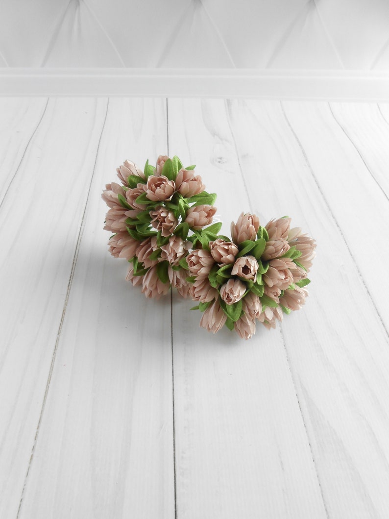 May include: Two clusters of artificial flowers with light pink petals and green leaves. The flowers are arranged in a round shape, and are set against a white wooden background.