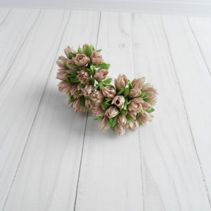 May include: Two bouquets of artificial tulips. The tulips are a soft pink color with green leaves. The bouquets are arranged in a circular shape. The background is a white wooden surface.