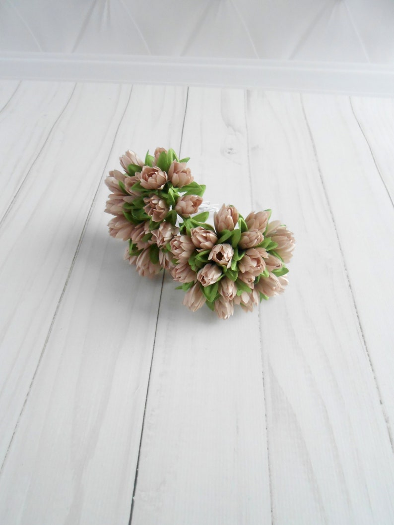 May include: Two bouquets of artificial tulips. The tulips are a soft pink color with green leaves. The bouquets are arranged in a circular shape. The background is a white wooden surface.
