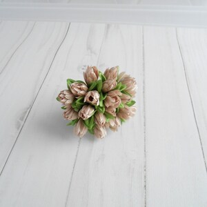 May include: A cluster of artificial tulips in shades of beige and light green. The faux flowers are arranged in a compact, rounded shape, with the petals closed. The arrangement sits on a white, wood-grain surface.