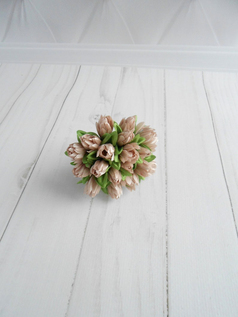 May include: A cluster of artificial tulips in shades of beige and light green. The faux flowers are arranged in a compact, rounded shape, with the petals closed. The arrangement sits on a white, wood-grain surface.