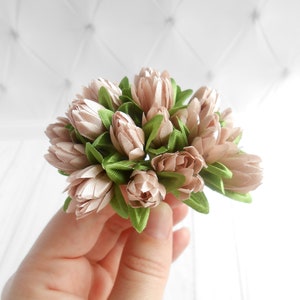 May include: A cluster of artificial flowers in shades of beige and green. The flower buds are beige, and the leaves are green. The flowers are held in a hand against a white background.