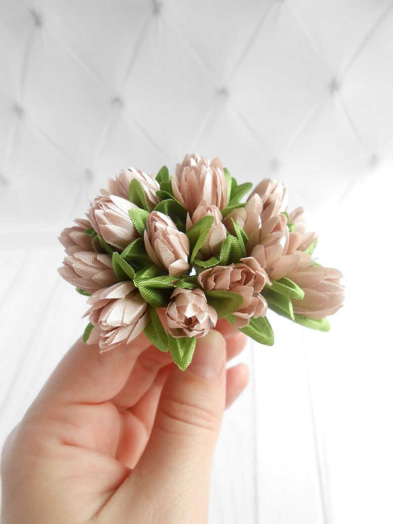 May include: A cluster of artificial flowers in shades of beige and green. The flower buds are beige, and the leaves are green. The flowers are held in a hand against a white background.