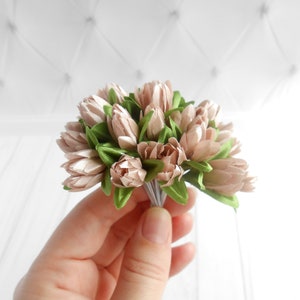 May include: A small bouquet of artificial flowers featuring light pink tulip-like blooms and green leaves. The flowers are tightly clustered and held together with a bundle of thin, silver-colored wires. The background is a white, textured surface.