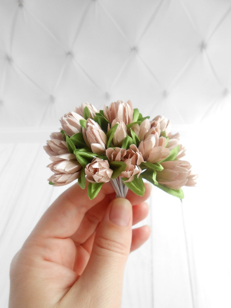 May include: A small bouquet of artificial flowers featuring light pink tulip-like blooms and green leaves. The flowers are tightly clustered and held together with a bundle of thin, silver-colored wires. The background is a white, textured surface.
