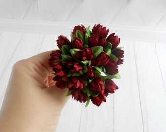 May include: A bouquet of burgundy artificial flowers with green leaves. The flowers are arranged in a round shape and are held in a hand.