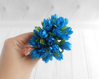 May include: A bouquet of blue silk flowers with green leaves. The flowers are arranged in a cluster and have a delicate, ruffled texture.